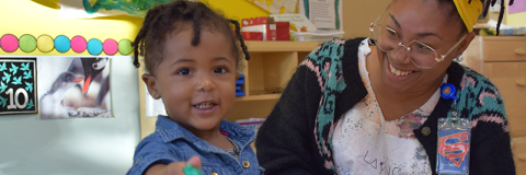 Smailing toddler points at viewer as mom looks on