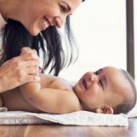 Happy mother playing with baby while changing his diaper. Smiling young woman with baby son on changing table at home. Close up of cheerful mom and toddler boy playing together. 