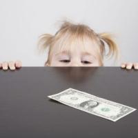 portrait of blonde caucasian baby nineteen month age with pigtails chubby face yellow shirt looking at dollar banknote on brown table 