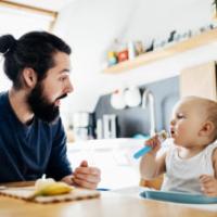 A single father sat at the kitchen table helping his baby eat some lunch. 