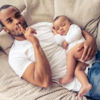 high angle view of handsome young afro american father looking at camera and showing silence sign while lying on sofa at home. little baby is sleeping in dad's arms 