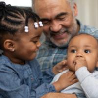 A senior aged ethnic grandfather is spending time with his young grandchildren. He is holding them and playing with them while sitting on a couch. 