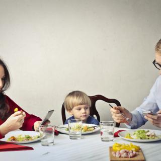 Family at dinner table with parents looking at cell phones while toddler is ignored. 