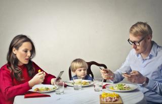 Family at dinner table with parents looking at cell phones while toddler is ignored. 