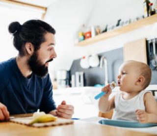 A single father sat at the kitchen table helping his baby eat some lunch.