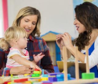 Mother and daughter work with preschool teacher. They are sitting at a table in the classroom working with educational toys. 