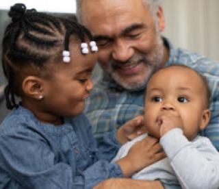 A senior aged ethnic grandfather is spending time with his young grandchildren. He is holding them and playing with them while sitting on a couch. 