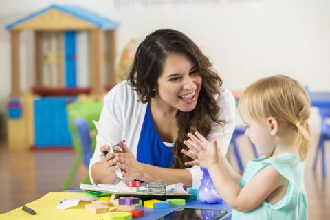 Cheerful female Hispanic teacher praises adorable blonde Caucasian little girl on accomplishing a project. The preschooler claps along with the teacher.